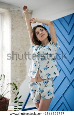 Young pretty slim brunette woman, wearing white pajamas,with blue flowers pattern, stretching in the morning. in light room near big window with white tulle and blue wall. Sleepwear design.