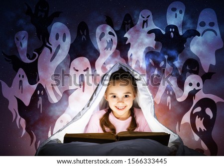 Cute girl reading kids book in bed. Child with flashlight hiding under blanket. Smiling girl in pajamas and funny ghosts silhouettes back on night sky. Child reading magic fairy tales. Happy childhood