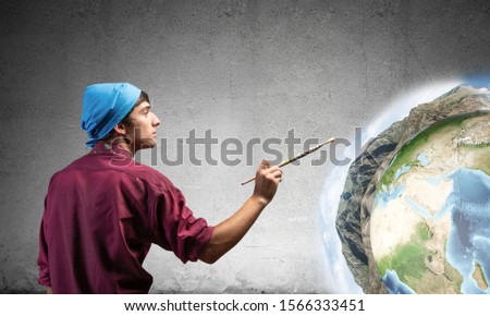 Young artist drawing earth globe. Male painter in dirty shirt and bandana with paintbrush on grey wall background. Creative hobby and artistic occupation. World ecology and environment concept.