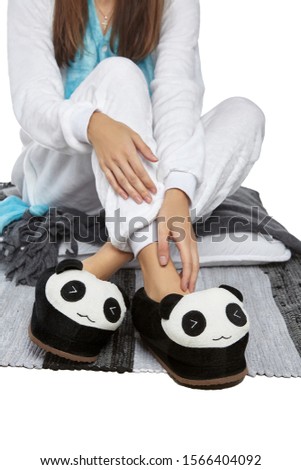Cropped medium shot of a girl in white velour pyjamas and black and white plush house slippers made in the form of panda with closed eyes. The girl is sitting on the striped carpet and a gray plaid.