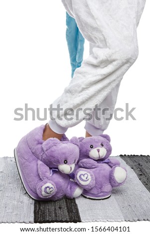 Close-up shot of female legs in white velour trousers and violet plush house slippers made in the form of a teddy bear with a silk ribbon. The girl is standing on the striped gray and white carpet.