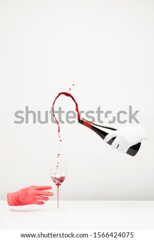 Surrealist picture of 2 hands in gloves on a white background and a glass of wine on a white table. One hand is holding a bottle of wine.