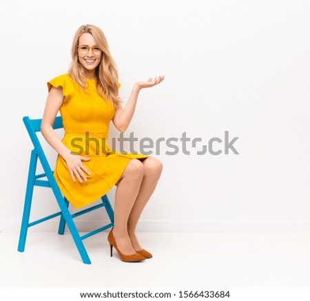 young pretty blonde woman feeling happy and smiling casually, looking to an object or concept held on the hand on the side against flat color wall