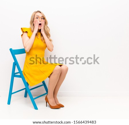 young pretty blonde woman feeling shocked and scared, looking terrified with open mouth and hands on cheeks against flat color wall