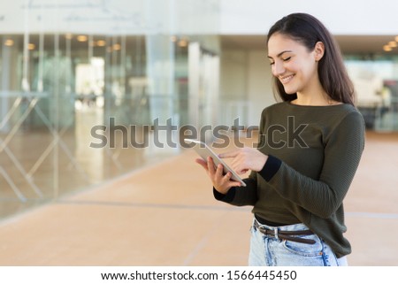 Happy cheerful student with tablet using Internet. Beautiful young woman in casual standing in hallway, holding digital device, touching screen, smiling. Internet connection concept