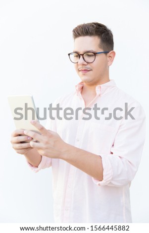 Focused nerdy guy in eyeglasses reading or watching content on tablet screen. Young man in glasses standing isolated over white background. Wi-fi concept