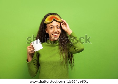 African-american young brunette woman's portrait in ski mask on green studio background. Concept of human emotions, facial expression, sales, ad, winter sport and holidays. Drinking tea or coffee.