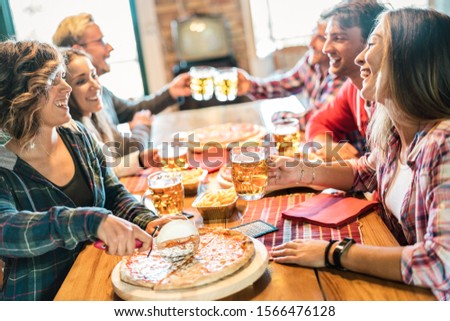 Young friends eating pizza at home on winter reunion - Friendship concept with happy people enjoying time together and having fun drinking brew pints - Cosy dinner place with focus on beer glass