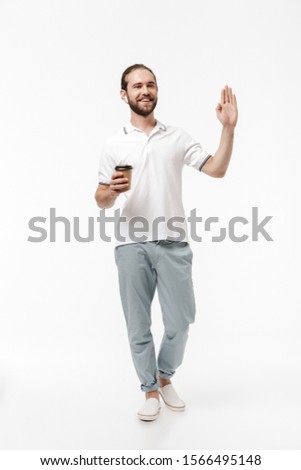 Photo of a young positive happy handsome bearded man walking isolated over white wall background listening music with earphones drinking coffee waving to someone.