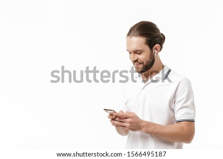 Photo of a young cheery positive happy handsome bearded man posing isolated over white wall background using mobile phone listening music with earphones.