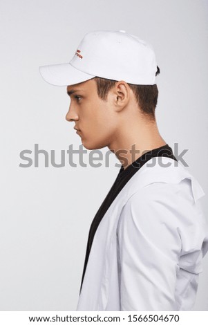 Cropped side photo of a dark-haired man, wearing white baseball cap with lettering "if you are not angry, you are not paying attention", black t-shirt and white shirt. He is posing on grey background 