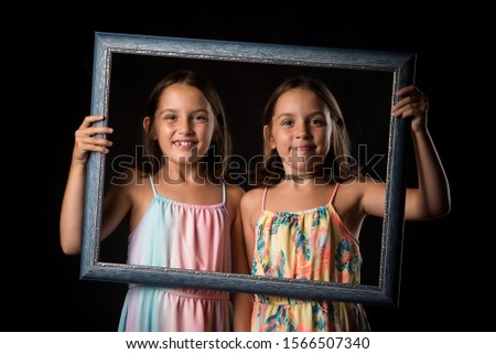 Identical twin girls are making happy expressions with picture frame. Children, sisters, girls posing in studio with picture frame, making different facial expressions. Family portrait, frontal view.