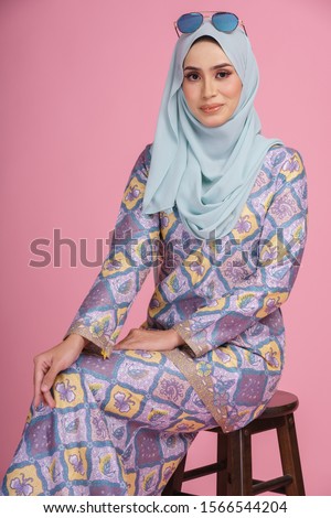 Beautiful female model wearing batik design "baju kurung" with light green hijab, sitting in various poses isolated over pink background. Eidul fitri fashion and beauty concept.