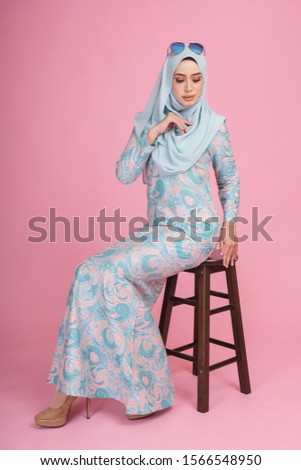 Beautiful female model wearing batik design "baju kurung" with light green hijab, sitting on a chair isolated over pink background. Eidul fitri fashion and beauty concept.