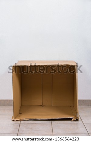 Open empty cardboard box on the floor inside a room with copy space on the wall above. Concept of improvised shelter of homeless and refugees.