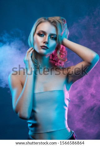 Beautiful blonde young girl with trendy makeup touches her face and hair with her hands, surrounded with blue and violet light in theatrical smoke. Clear, healthy skin. Advertising, commercial design