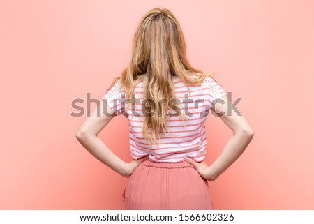 young pretty blonde woman feeling confused or full or doubts and questions, wondering, with hands on hips, rear view against flat color wall