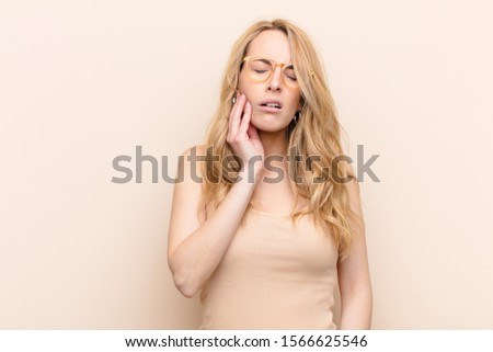 young pretty blonde woman holding cheek and suffering painful toothache, feeling ill, miserable and unhappy, looking for a dentist against flat color wall
