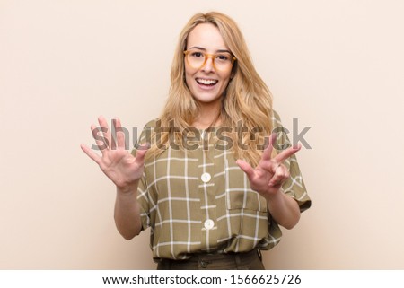 young pretty blonde woman smiling and looking friendly, showing number eight or eighth with hand forward, counting down against flat color wall