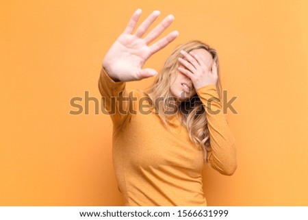 young pretty blonde woman covering face with hand and putting other hand up front to stop camera, refusing photos or pictures against flat color wall