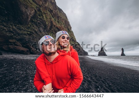 A guy and a girl in red sweaters and gray hats are hugging on a black beach, next to Vic. Happy young couple in a hat with a flag. A happy stylish smiling couple walking and kissing in Iceland.