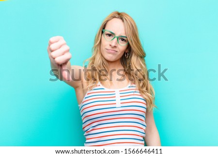 young pretty blonde woman feeling cross, angry, annoyed, disappointed or displeased, showing thumbs down with a serious look against flat color wall