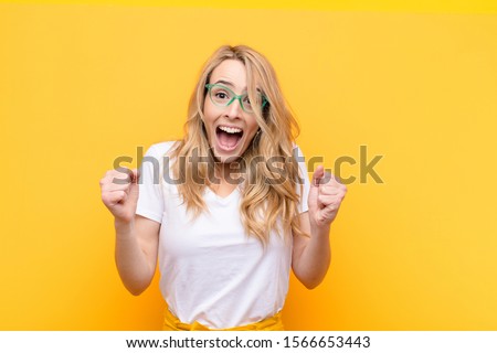 young pretty blonde woman feeling shocked, excited and happy, laughing and celebrating success, saying wow! against flat color wall