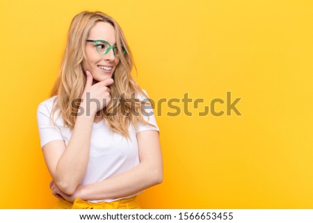 young pretty blonde woman smiling with a happy, confident expression with hand on chin, wondering and looking to the side against flat color wall