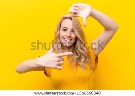 young pretty blonde woman feeling happy, friendly and positive, smiling and making a portrait or photo frame with hands against flat color wall