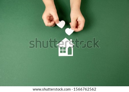 Child hands holding white paper house and heart on green background. Family home and real estate concept. Flat lay, copy space