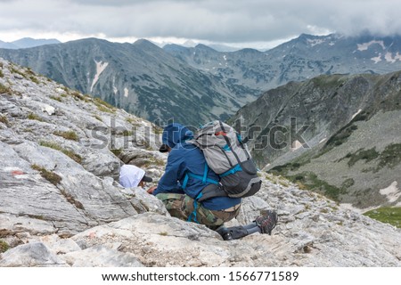 Man helps to injured, wounded young boy, who lying among rocks in a mountain. Majestic peaks at background. Selective focus. First aid, survival concept, help someone in need. Royalty-Free Stock Photo #1566771589