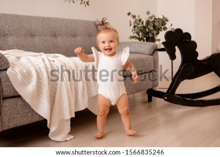 healthy baby girl in a white bodysuit in a room next to a gray sofa is learning to walk. the comfort of the home. the child smiles