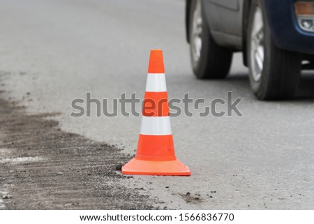 Warning traffic road cone standing on street asphalt city road during roadworks, road repairing, asphalt pavement works on highway. Unrecognizable auto driving along road in blurred background.