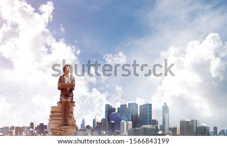 Smiling woman holding open and looking up. Beautiful girl in business suit sitting on stack of big books. Businesswoman with textbook on background of cityscape panorama. Knowledge and education