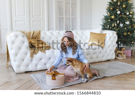 Beautiful young curly haired girl with her pet, dog and friend, hugging and considering Christmas gifts on the background of Christmas tree, garlands, fireplace and bokeh in a cozy white living room.