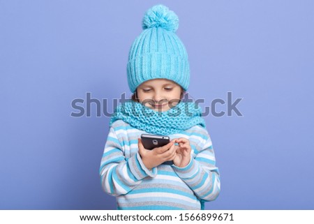 Cute preschooler little girl standing isolated over blue studio background, child using modern smartphone, smart small kid surfing internet watch cartoon on cellphone, wearing warm winter clothing.