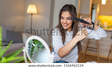 Woman straightening hair with straightener. Beautiful woman straightening her hair. Beautiful young smiling woman using a hair straightener while looking into the mirror Royalty-Free Stock Photo #1566962134