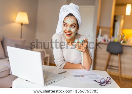 Woman wearing facial mask enjoying at home and using her laptop. Multitasking young woman with beauty treatment facial mask chatting using smartphone in livingroom