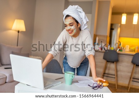 Woman wearing facial mask enjoying at home and using her laptop. Multitasking young woman with beauty treatment facial mask chatting using smartphone in livingroom