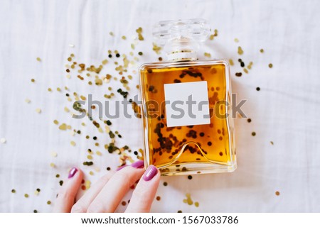 perfume bottle next to gold sequins on a white sheets and a female hand