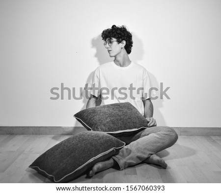 black and white photography.young handsome Italian teenager model boy posing for fashion photo shooting,sitting on wooden floor and playing with pillows during pillow fight.white background