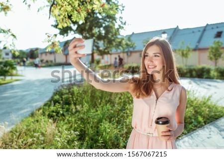 girl in pink dress, summer city park pictures on smartphone, selfie, holds cup coffee tea, smiles happy, video call, online application Internet. Modern fashionable beautiful woman. Tanned long hair.