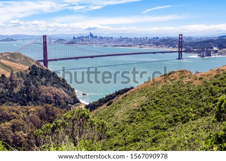 Picture of the Golden Gate Bridge with San Francisco in the background.  Green rolling mountains in the foreground with aqua water flowing under the bridge.