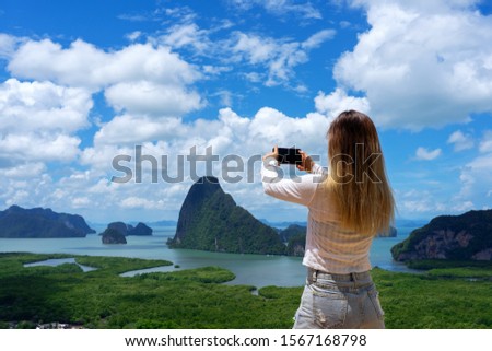 Asian woman holding smart phone taking photo of small islands and sea with green mangrove forest and blue sky background in Phang Nga Bay, Thailand.