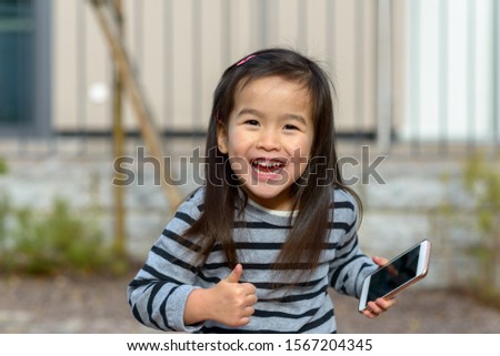 Vivacious playful little Asian girl giving a thumbs up with a delightful smile as she holds a mobile phone outdoors