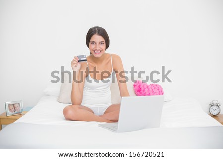 Pleased young brown haired model in white pajamas shopping online with her laptop in bright bedroom