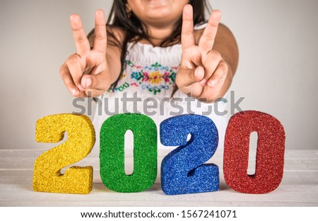 Young girl making peace sign above bright and colorful 2020 3d numbers - new years celebrations concept image.