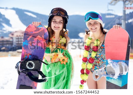 Two young female snowboarders in colorful clothing, swimsuit, bra, skirt and flower wreath posing with snowboards on background of blue sky and winter mountain resort. Sports and festivity concept.