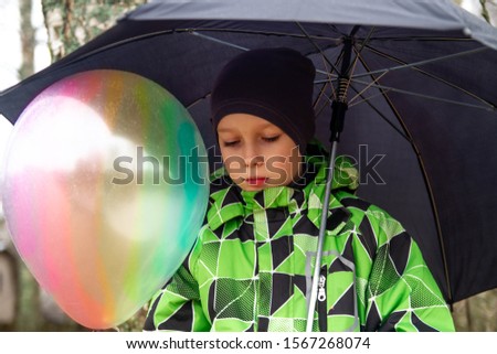 The sad boy was cold and wet from the rain. A boy with a multicolored balloon took shelter from the rain under a black umbrella. The balloon is like a rainbow.