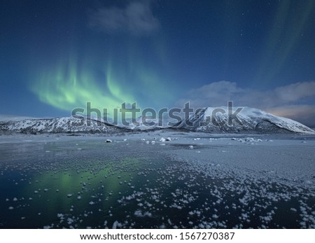 A beautiful view of the half froze lake surrounded by snow covered hills under the northern lights